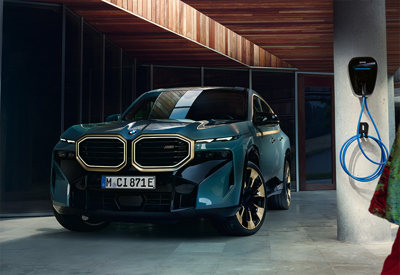 Der BMW XM Plug-In-Hybrid (PHEV) präsentiert sich als kraftvolles, modernes SUV mit markanter Designsprache und sportlichen Akzenten. Die breite Front ist geprägt von einer großen, vertikalen BMW-Niere, flankiert von schmalen LED-Scheinwerfern und Lufteinlässen. Im Bild ist eine Wallbox, die an einer zu Rundstütze befestigt ist, zu erkennen.