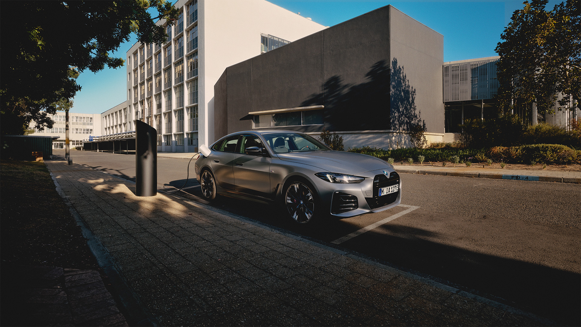 Die Front des BMW i4 (2024) präsentiert sich mit einem modernen, dynamischen und elektrifizierten Design. Die großzügige, geschlossene BMW-Niere in hochglänzendem Schwarz dominiert das Erscheinungsbild und ist ein unverkennbares Merkmal der BMW i-Modelle. Die schmalen LED-Scheinwerfer mit moderner Lichtsignatur und das sportlich gestaltete Frontspoiler-Design unterstreichen die fortschrittliche, technisch hochwertige Optik. Die Frontschürze mit aerodynamisch optimierten Lufteinlässen und eleganten Akzenten sorgt für eine klare, kraftvolle Präsenz. Insgesamt vermittelt die Front des BMW i4 (2024) eine exklusive, zukunftsorientierte und sportliche Erscheinung. Das Fahrzeug steht an der Straßenseite auf einer markierten Parkfläche. Der vollelektrische BMW i4 wird an einer zylinderförmigen Ladesäule geladen. Im Hintergrund ist eine rationalistische Architektur zu sehen.