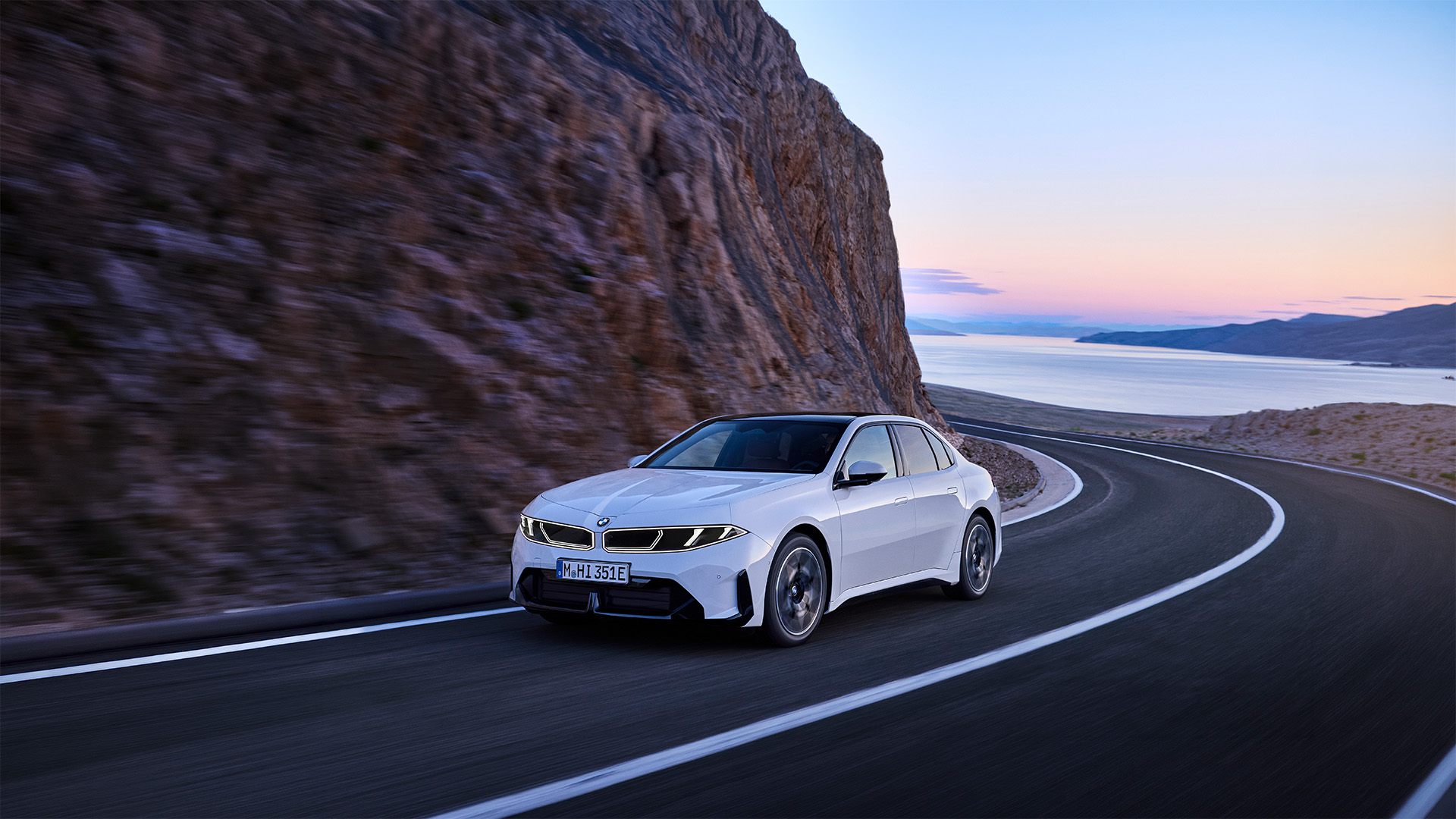 BMW i3 Limousine First Edition in Alpinweiß während der Fahrt auf eine Straße zwischen einem See und Felsen