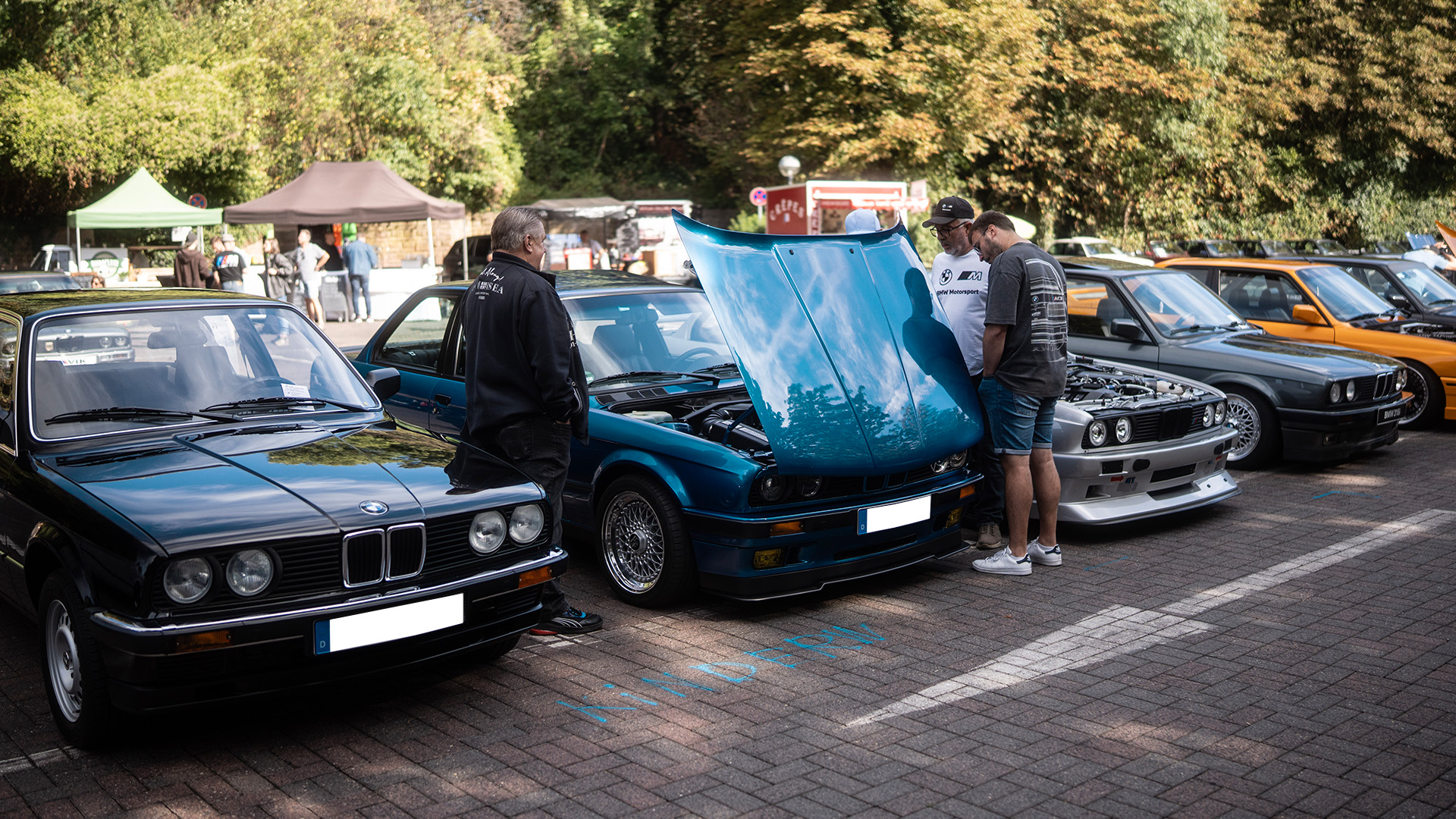 Besucher auf dem Burgparkplatz schauen sich BMW E30 Technik unter der Motorhaube an.